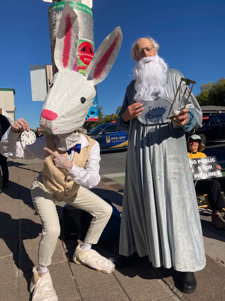 Two people in costume at a street demonstration: a white rabbit holding a pocket watch, and Father Time with a long white beard and an hourglass.