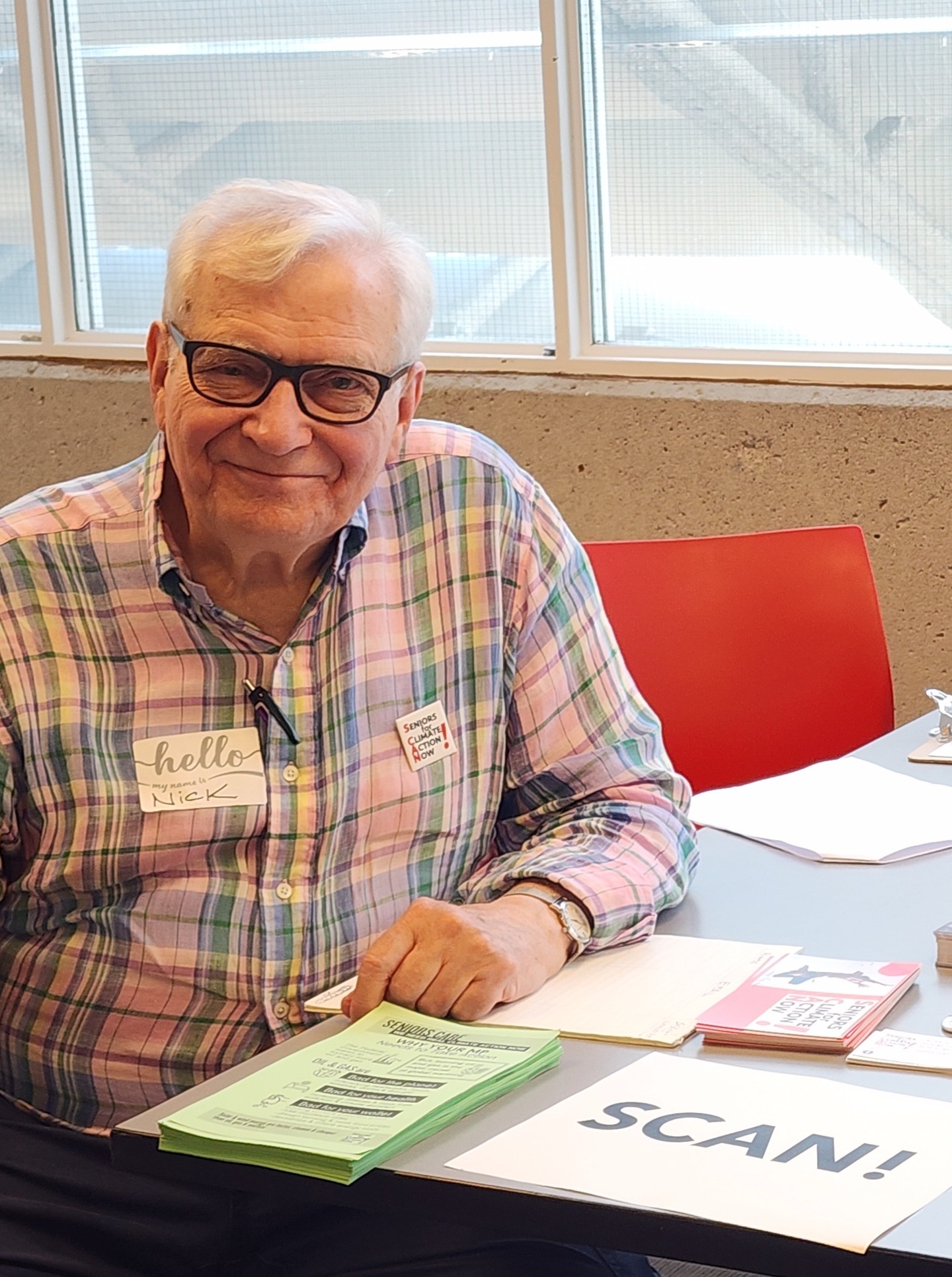 A smiling older man in glasses and a plaid shirt, with a name tag, sits at a welcome table with leaflets.