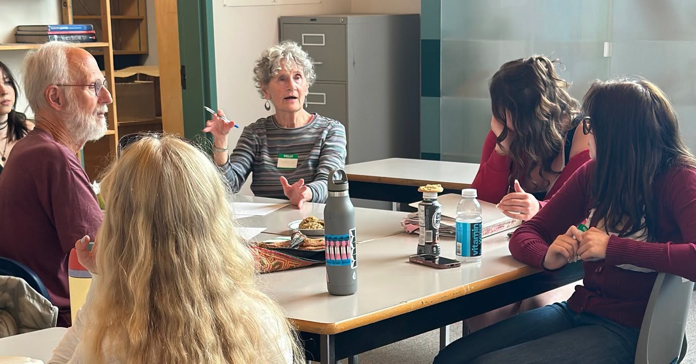 Une femme âgée aux cheveux gris bouclés parle en gesticulant à une table, pendant qu’un homme à la barbe grise et des personnes plus jeunes l’écoutent, lors d’un café climat d’Aînés pour le climat.