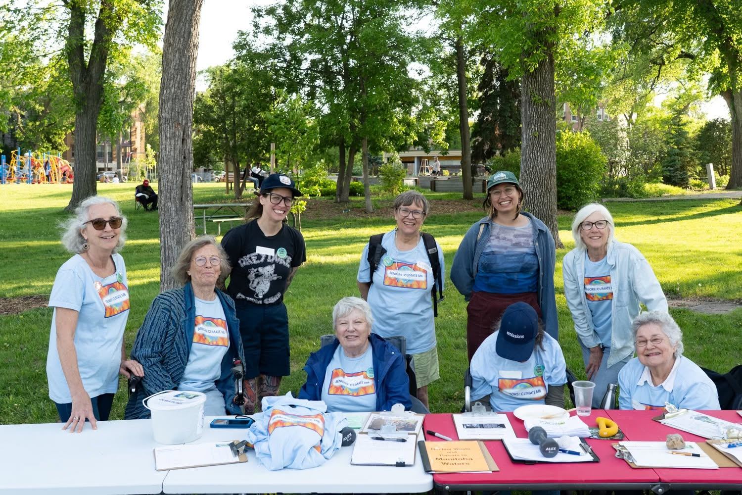 Des membres souriants, en t-shirts d’Aînés pour le climat, assis et debout autour d’une table d’information dans un parc.