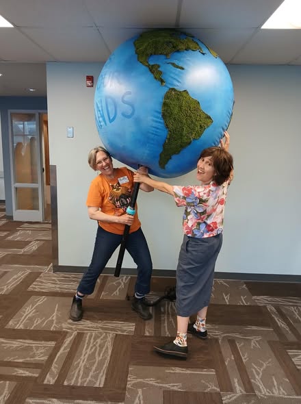 Deux femmes, dont l’une en t-shirt orange, tiennent un grand globe gonflable au-dessus de leur tête, dans un couloir lumineux.