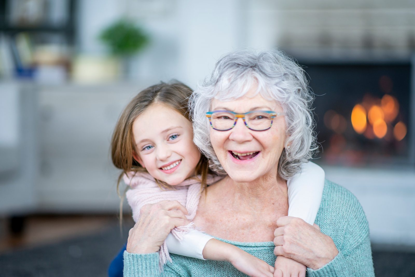 Une grand-mère rieuse aux lunettes colorées se fait serrer dans les bras par sa petite-fille souriante, dans un salon devant un feu de foyer.